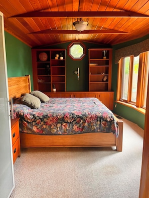 Ein Schlafzimmer mit Holzbalkendecke, einem Bett mit floralem Überwurf, integrierten Regalen und einem Fenster mit Vorhängen.