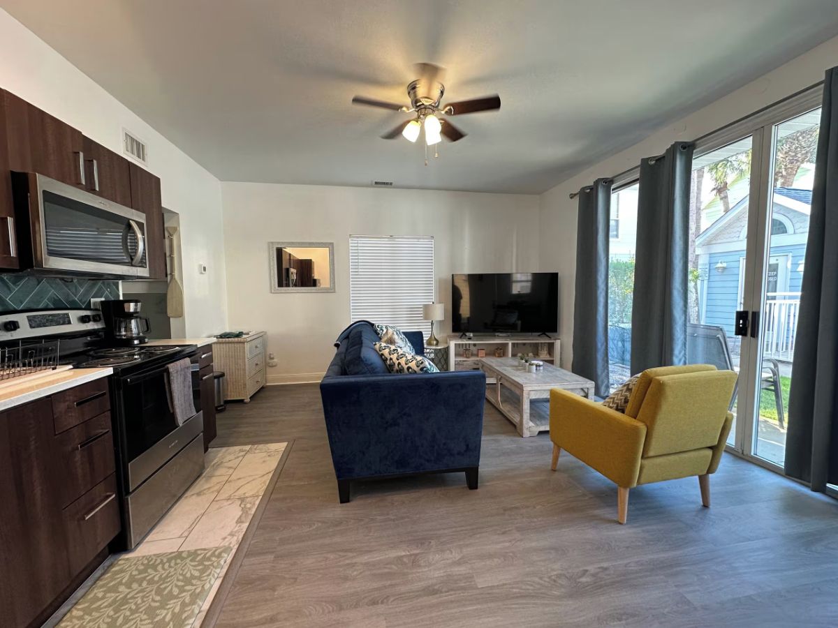 A modern kitchen with dark cabinetry, a stove, and a microwave. There is a blue sofa and a yellow armchair in the living area, with a TV and a coffee table.