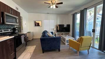 A modern kitchen with dark cabinetry, a stove, and a microwave. There is a blue sofa and a yellow armchair in the living area, with a TV and a coffee table.