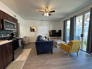 A modern kitchen with dark cabinetry, a stove, and a microwave. There is a blue sofa and a yellow armchair in the living area, with a TV and a coffee table. - Cozy Beach Retreat - Pool - Steps to Beach - Ground Condo - sleeps 6 (Destin)