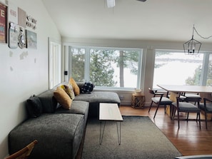 A modern living room with a grey sectional sofa, a small white table, and a dining area with wooden chairs. Large windows provide a view of a lake and trees. - Charming Lakeside Cottage (North Bay)