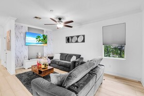 A modern living room with a grey sofa, a coffee table with decorative items, a flat-screen TV mounted on the wall, and a ceiling fan. - Riverfront 5BR Villa - Private Dock & Pool (Fort Myers)