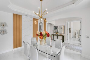 A modern dining area with a glass table, white chairs, a chandelier, and a vase with flowers. - Riverfront 5BR Villa - Private Dock & Pool (Fort Myers)