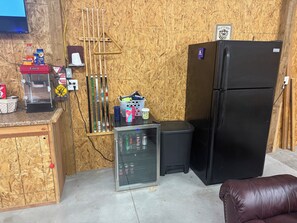 A compact room with a black refrigerator, a small glass cabinet with beverages, a trash bin, and a wooden wall with a mounted television. -  Shed in Great Bend perfect for your next fun gathering. (Great Bend)
