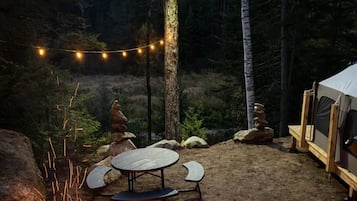 Un terrain de camping équipé d’un foyer, d’une table avec des bancs, de guirlandes lumineuses et d’une tente.