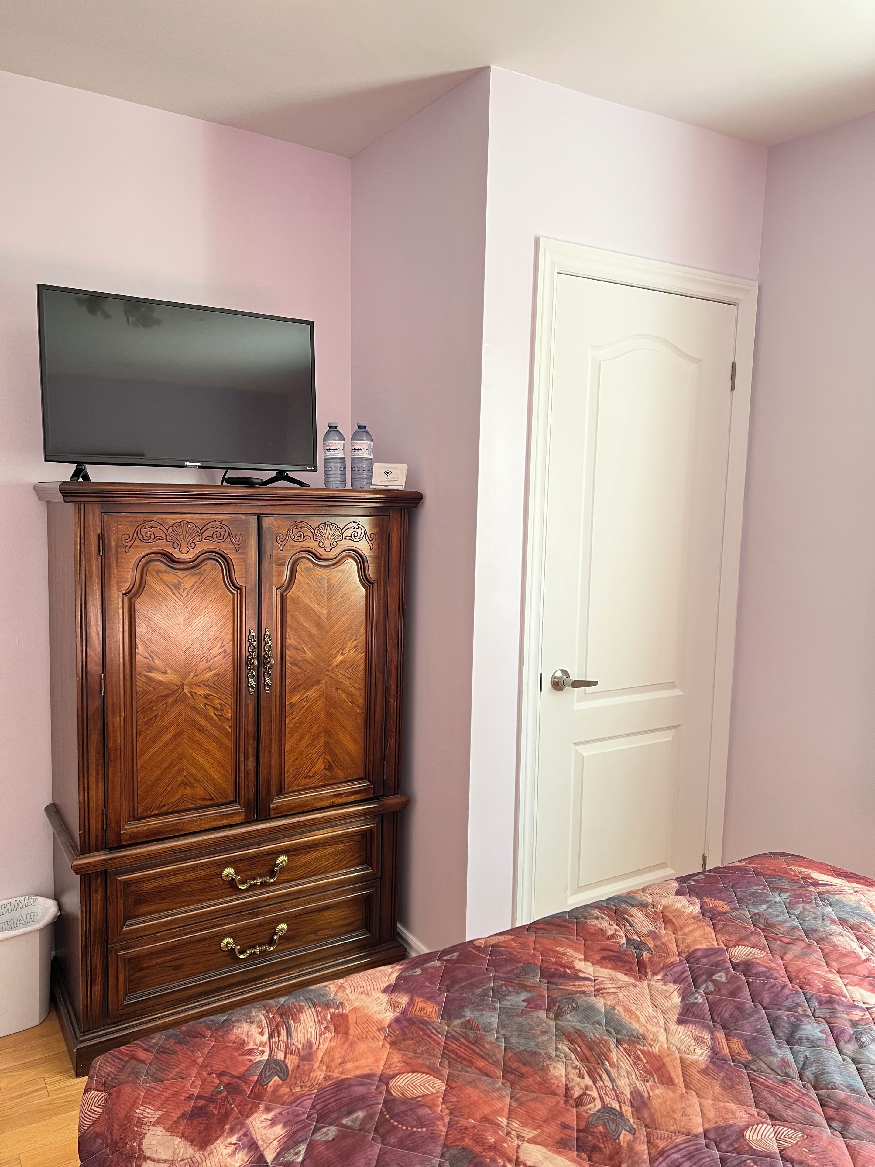 A bedroom with a wooden dresser, a television on top, a door, and a bed with a patterned bedspread.