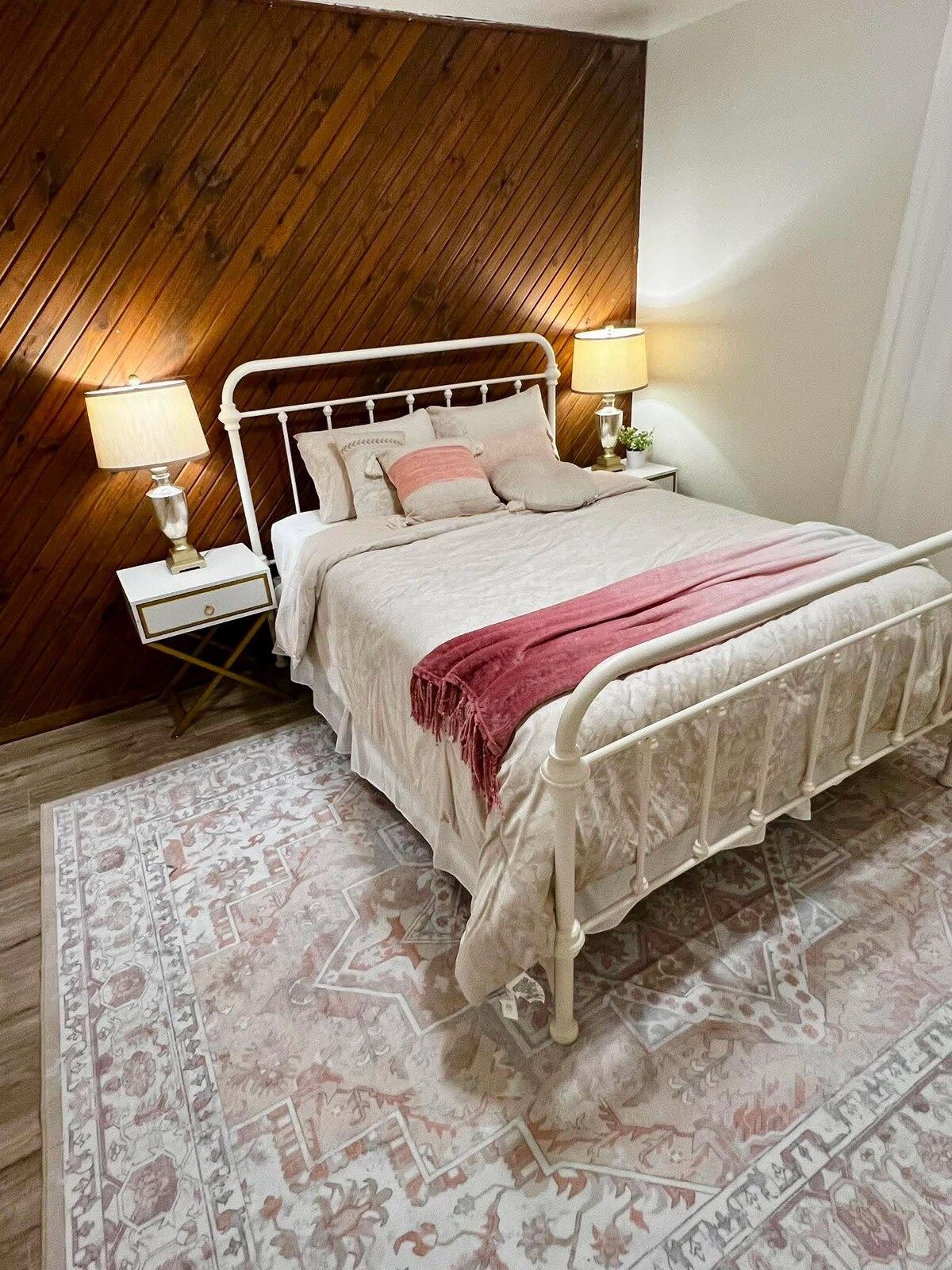 A bedroom with a wooden sloped ceiling, a white metal bed frame, two bedside lamps, a patterned rug, and a pink throw blanket.