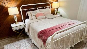 A bedroom with a wooden sloped ceiling, a white metal bed frame, two bedside lamps, a patterned rug, and a pink throw blanket.