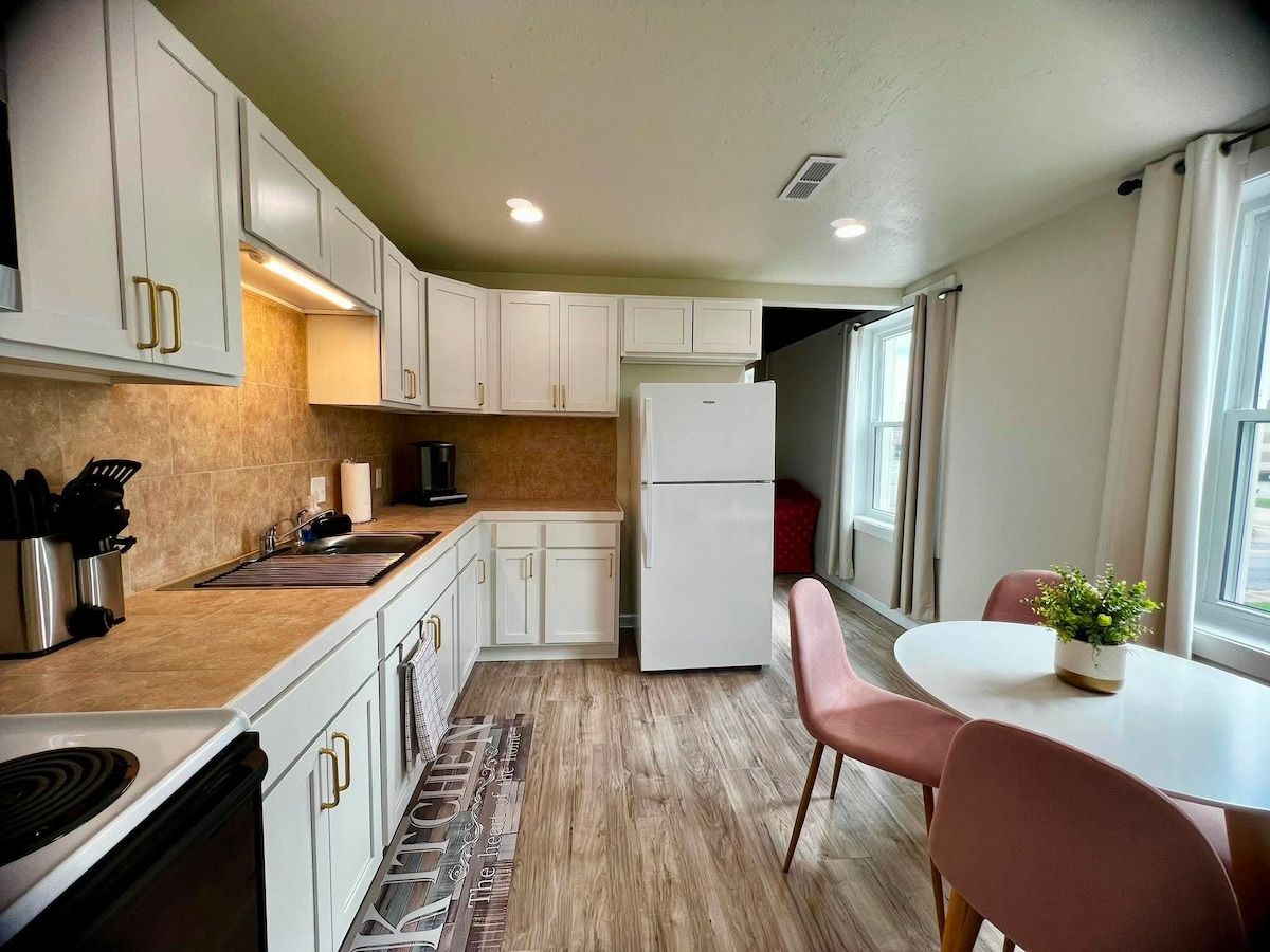 A compact kitchen with white cabinets, a refrigerator, a dining table with chairs, and a window providing natural light.
