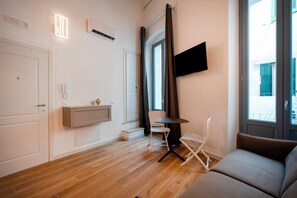 A modern living room with a sofa, a small table, a chair, a mounted TV, and a window with curtains. - Palazzo Dell'Erba - Deluxe (Monopoli)