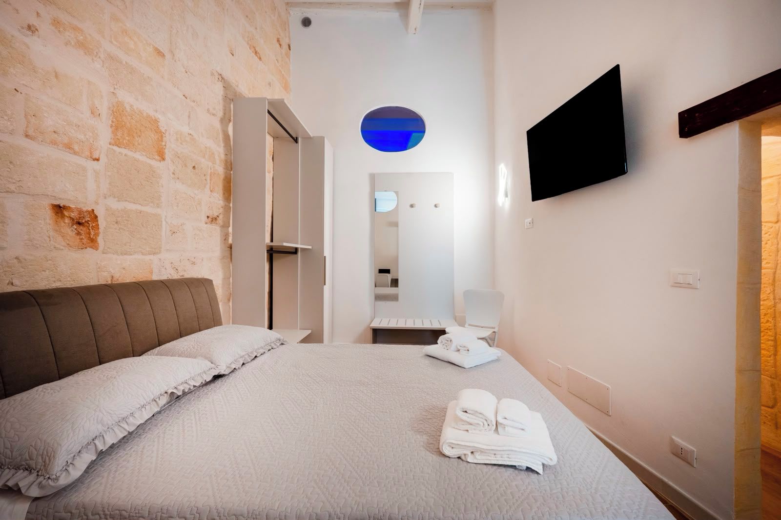 A hotel room with a bed, a flat-screen TV, a mirror, and a towel rack.