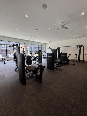 Un gimnasio con diversas máquinas de ejercicio, amplias ventanas y ventiladores de techo.