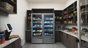 A self-service beverage station with refrigerated drink dispensers and a variety of snacks and condiments on shelves. - Landing | Spectacular 1BD in Lochridge (Cary)