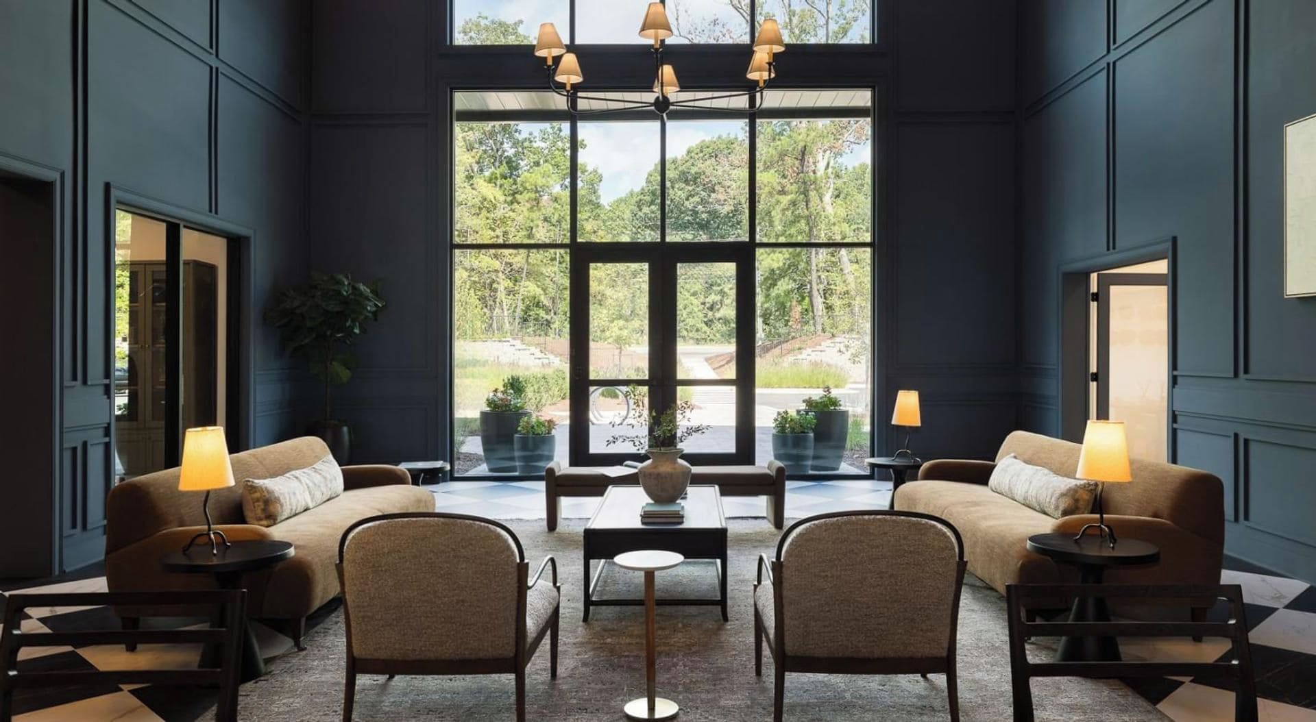 A modern interior with two sofas, a coffee table, and chairs arranged around it. Large windows provide a view of greenery outside.