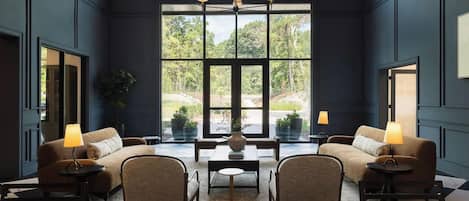 A modern interior with two sofas, a coffee table, and chairs arranged around it. Large windows provide a view of greenery outside.