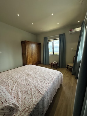 Un dormitorio con una cama grande, un armario de madera, una mesita, una silla y una ventana con cortinas.