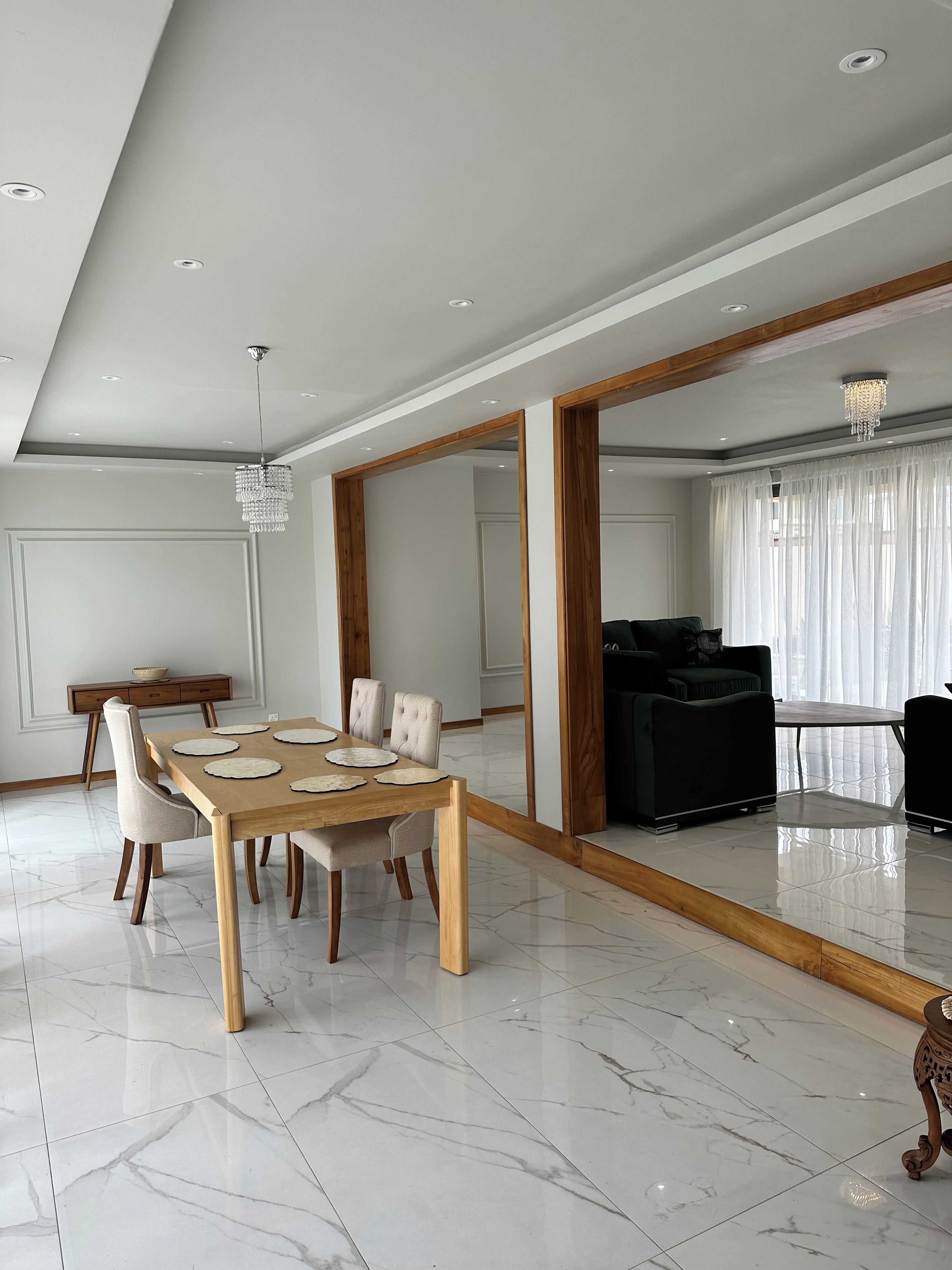 Un espace de salle à manger moderne, avec une table et des chaises en bois, un lustre et un grand miroir qui reflète la pièce.