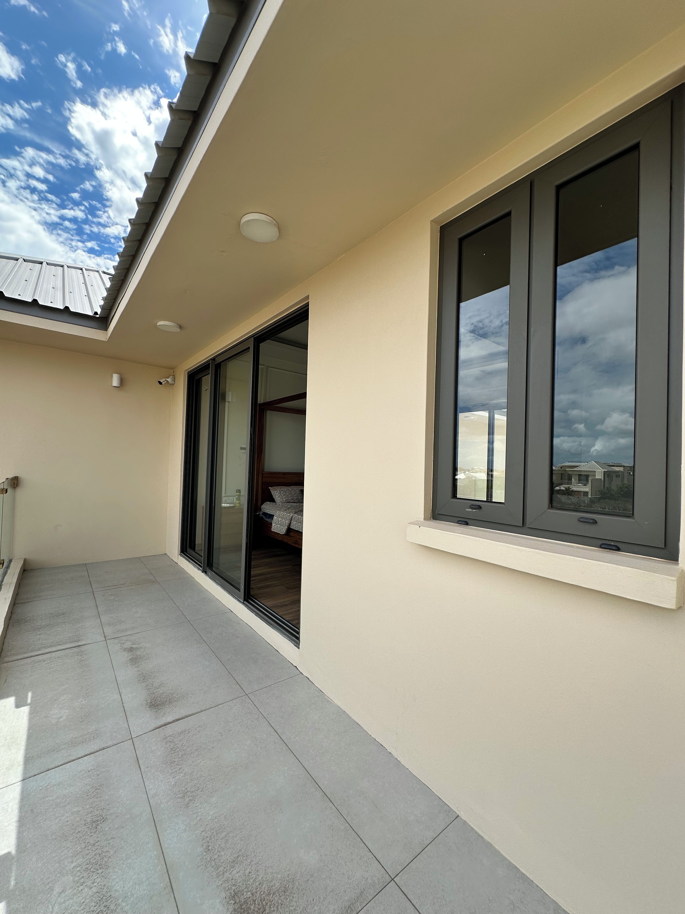 Un balcone con pavimento piastrellato, una porta scorrevole in vetro che dà su una camera da letto e un'ampia finestra con vista sul cielo.