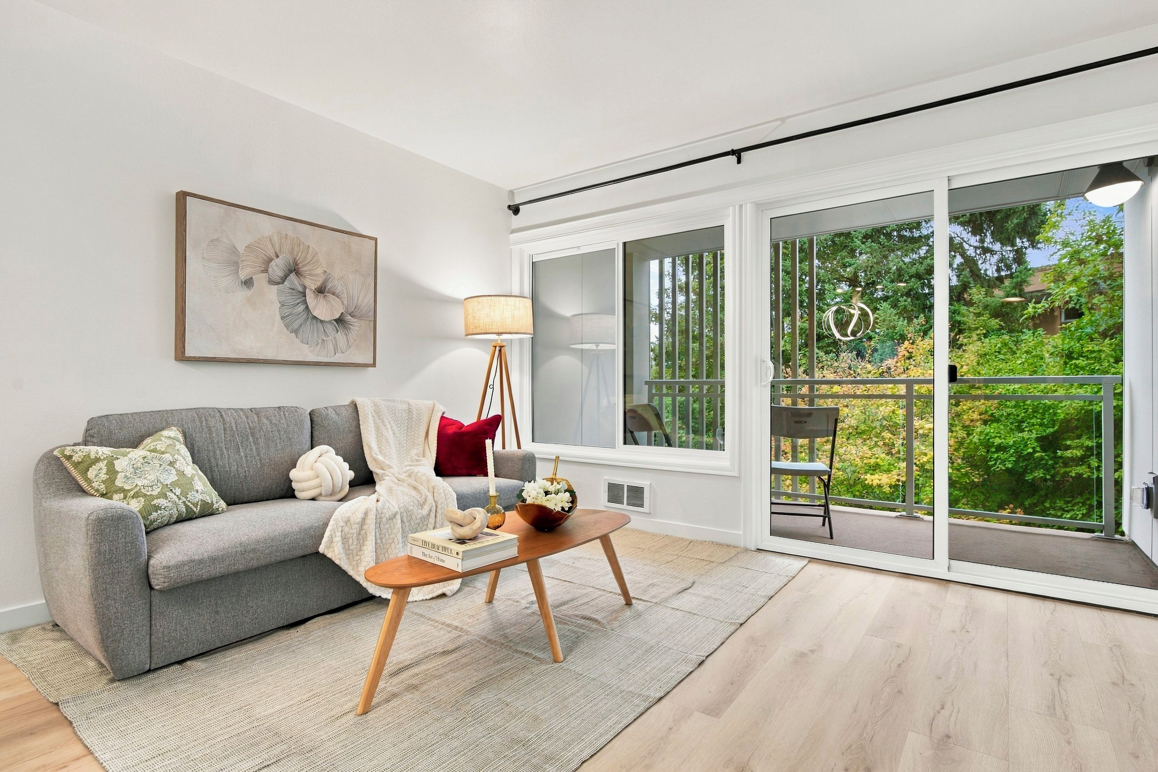 A living room with a grey sofa, a floor lamp, a coffee table, and a large window with a view of greenery.