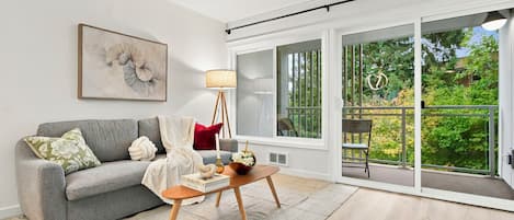 A living room with a grey sofa, a floor lamp, a coffee table, and a large window with a view of greenery.