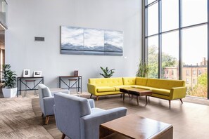 Un salon moderne avec un canapé jaune, un fauteuil gris et une table basse en bois. Une grande fenêtre offre une vue sur les bâtiments et les arbres.