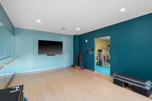 A spacious room with teal walls, a mounted TV, a wooden floor, and a doorway leading to a gym area with exercise equipment. - Landing | Stylish 1BD in Central West End (St. Louis)