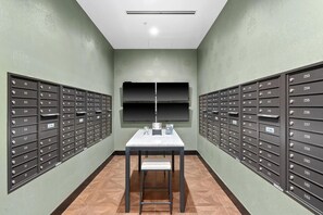 A row of mailboxes with numbers, a table with a chair, and a wall-mounted shelf. - Landing | Chic 2BD in Central West End (St. Louis)