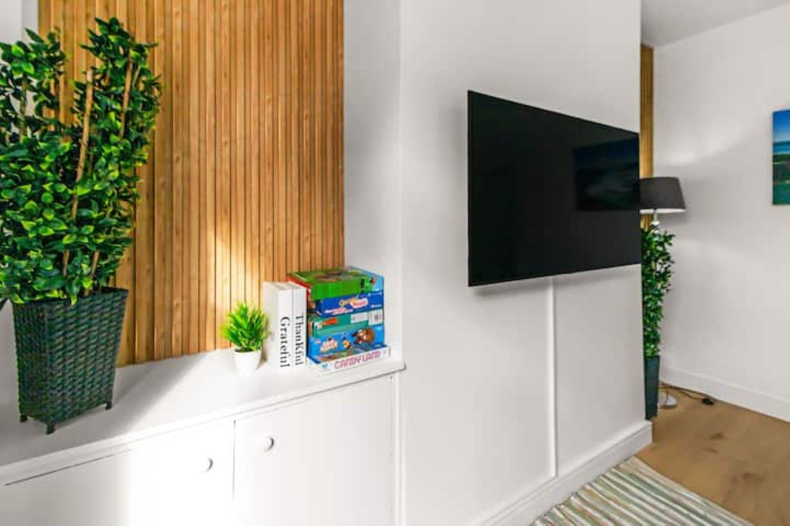 Un salon moderne avec un téléviseur à écran plat fixé au mur, un mur recouvert de panneaux en bois, une plante en pot, une petite table sur laquelle se trouve une boîte de jeux et un tapis à rayures.
