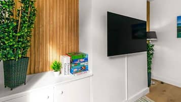 Un salon moderne avec un téléviseur à écran plat fixé au mur, un mur recouvert de panneaux en bois, une plante en pot, une petite table sur laquelle se trouve une boîte de jeux et un tapis à rayures.