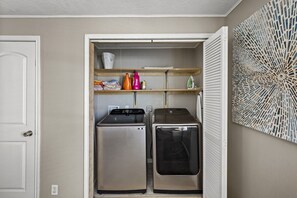 Une buanderie équipée de deux machines à laver et deux sèche-linge superposés, d’étagères où sont pliés les vêtements et d’un élément décoratif mural.