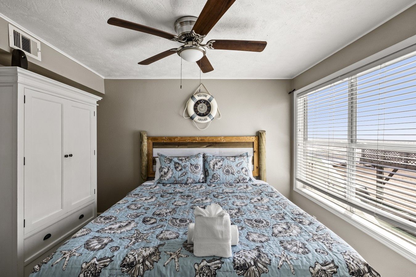A bedroom with a bed, a ceiling fan, a window with blinds, and a wall-mounted clock.