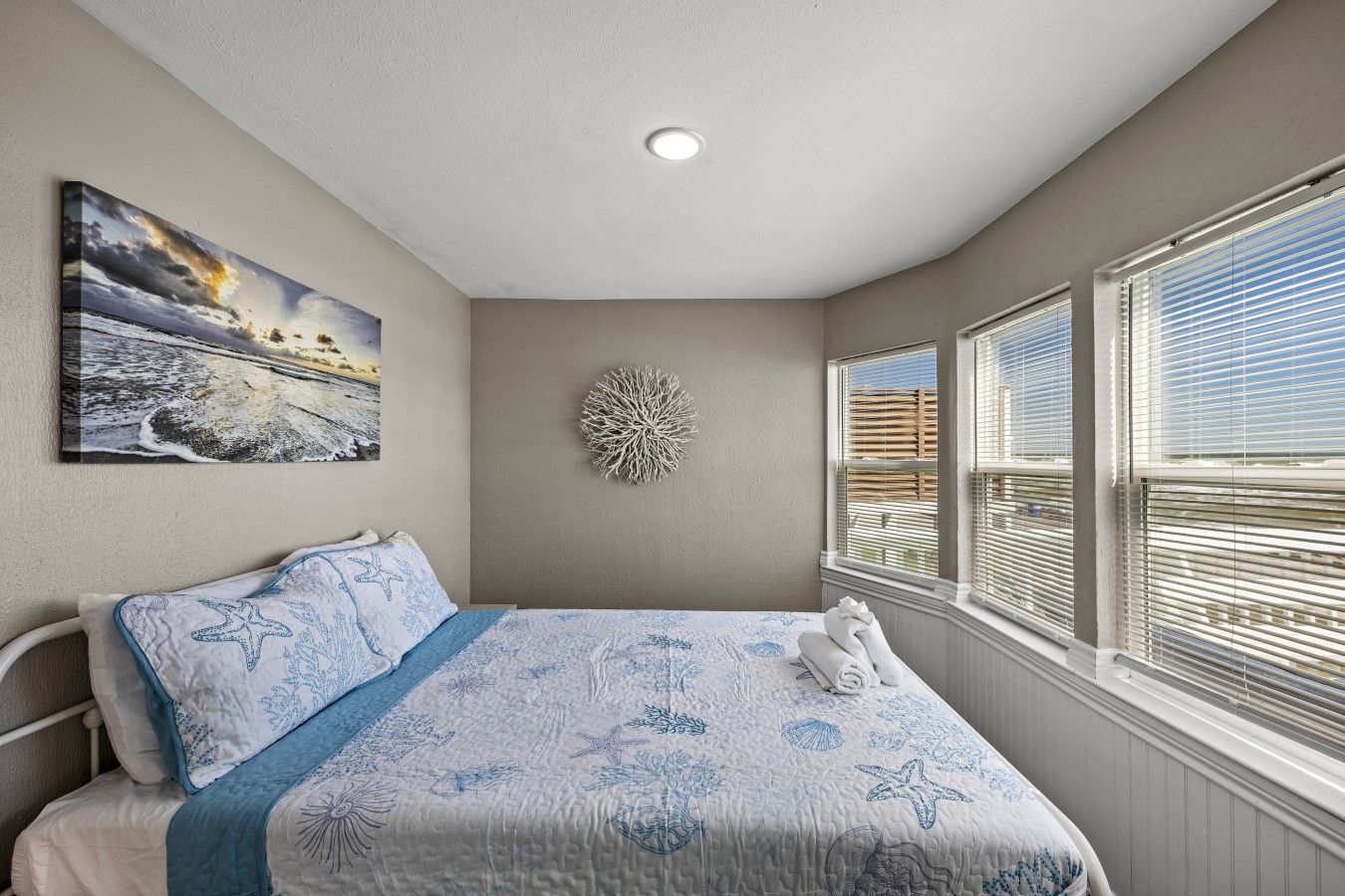 A bedroom with a bed, a wall-mounted painting, a decorative coral piece, and a window with blinds.
