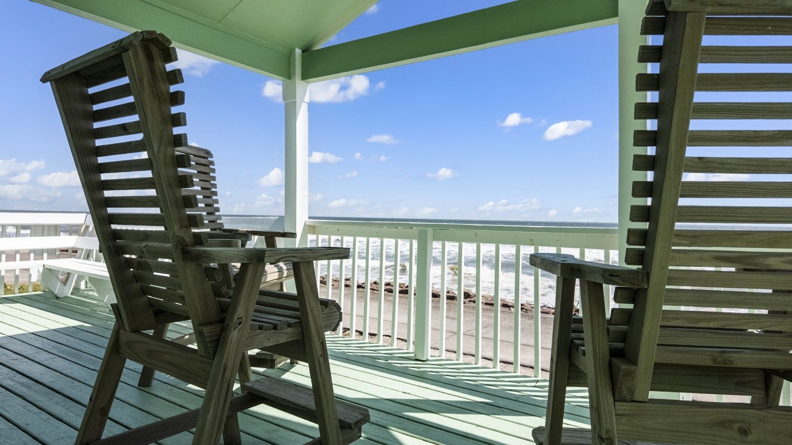 A wooden deck with two chairs and a bench, overlooking a beach with a clear sky.