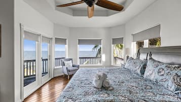 A bedroom with a large bed, a ceiling fan, and a view of the ocean through multiple windows.