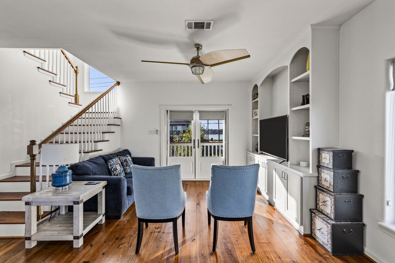 A spacious living room with a staircase, a ceiling fan, a television, and a view through a door.