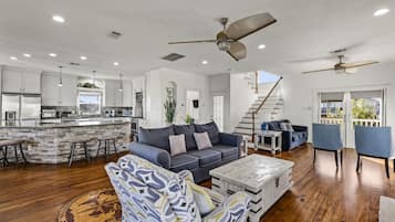 A spacious living area with a sofa, coffee table, and chairs, a kitchen with white cabinets, and a staircase leading to a balcony.