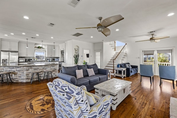 A spacious living area with a sofa, coffee table, and chairs, a kitchen with white cabinets, and a staircase leading to a balcony. - Far Harbor- Waterfront | Close to Kemah Boardwalk (Kemah Area)