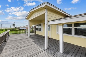 A covered outdoor deck with a wooden floor, white railings, and a yellow building facade. - Warblers Roost- Bird Watcher Paradise | Great view (Crystal Beach)