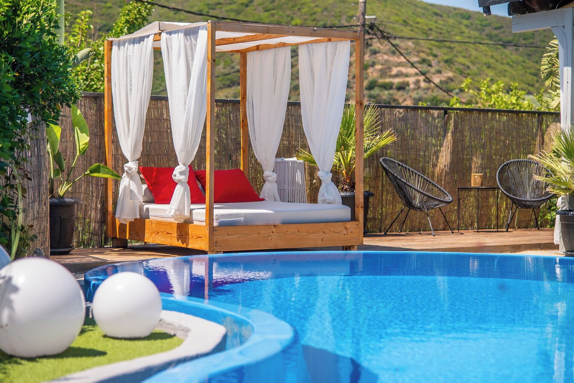 A wooden four-poster bed with white curtains, red pillows, and a small statue, adjacent to a swimming pool with blue water.