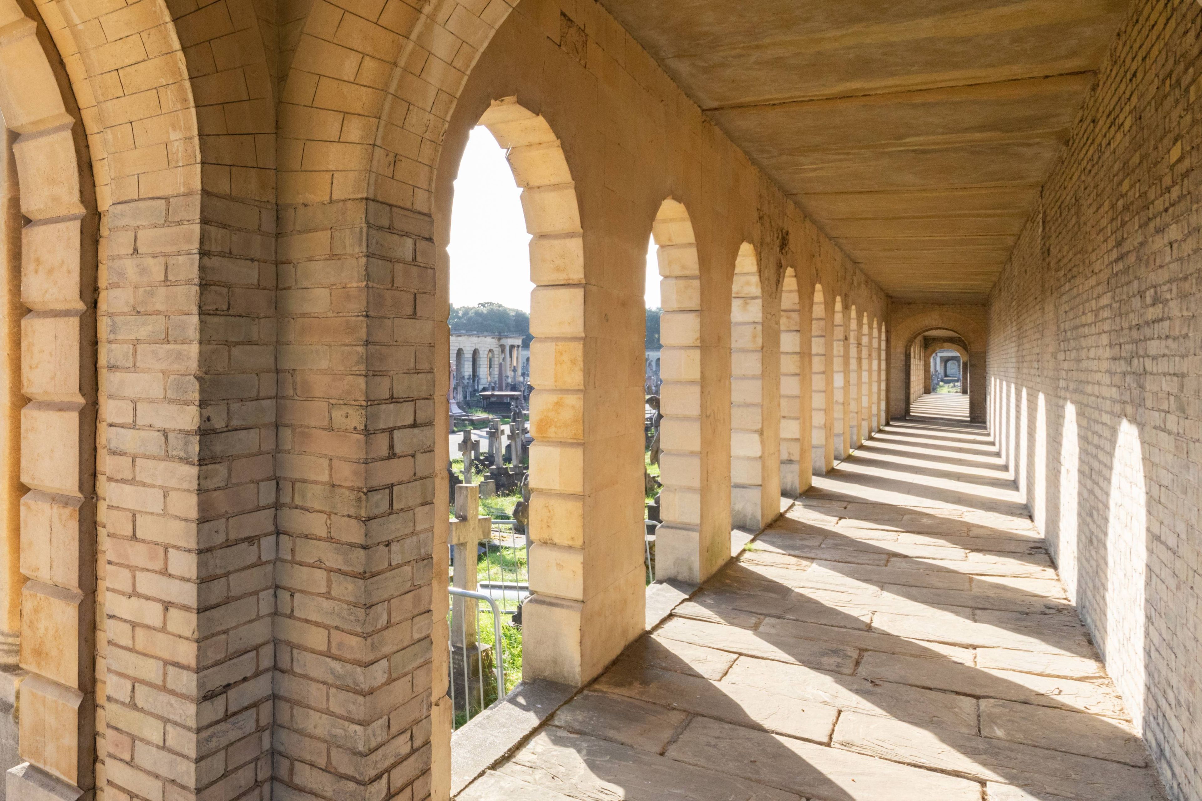 A corridor with stone arches and a tiled floor, leading to a view of a bridge.