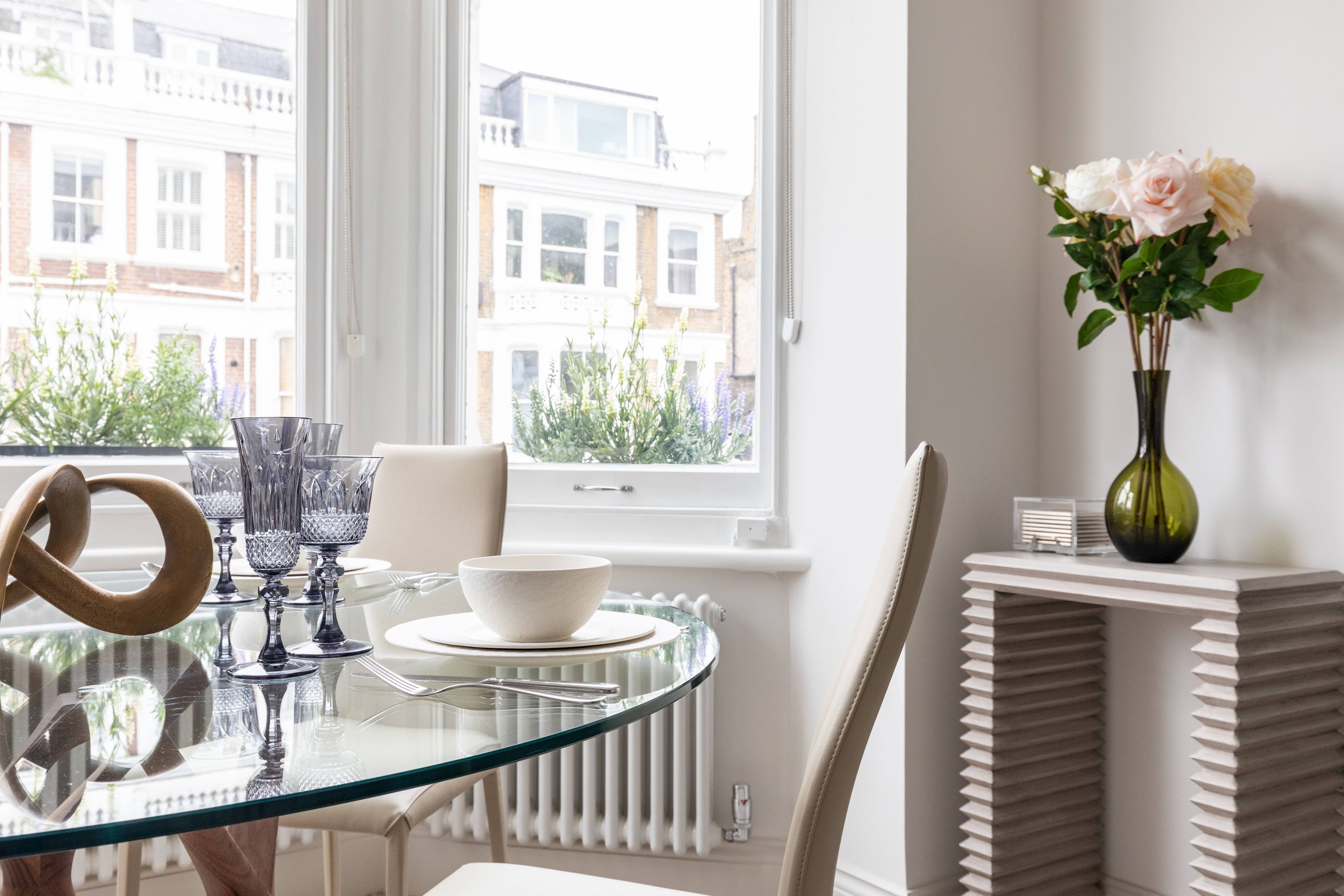 A dining area with a glass table, white plates, and cutlery. There is a vase with flowers on a white console table.