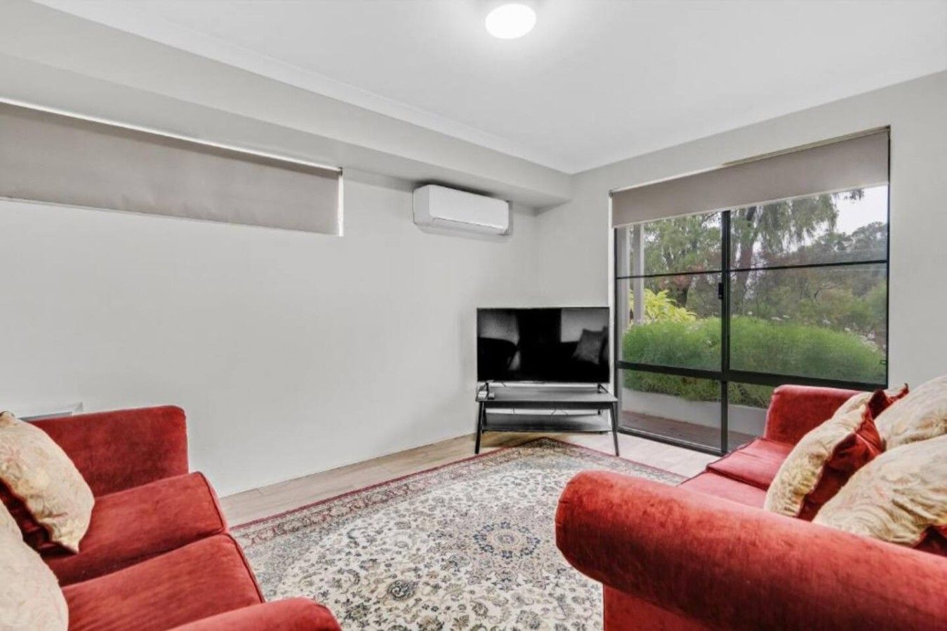 A modern living room with a flat-screen TV, red sofas, a patterned rug, and a large window with a view of greenery.