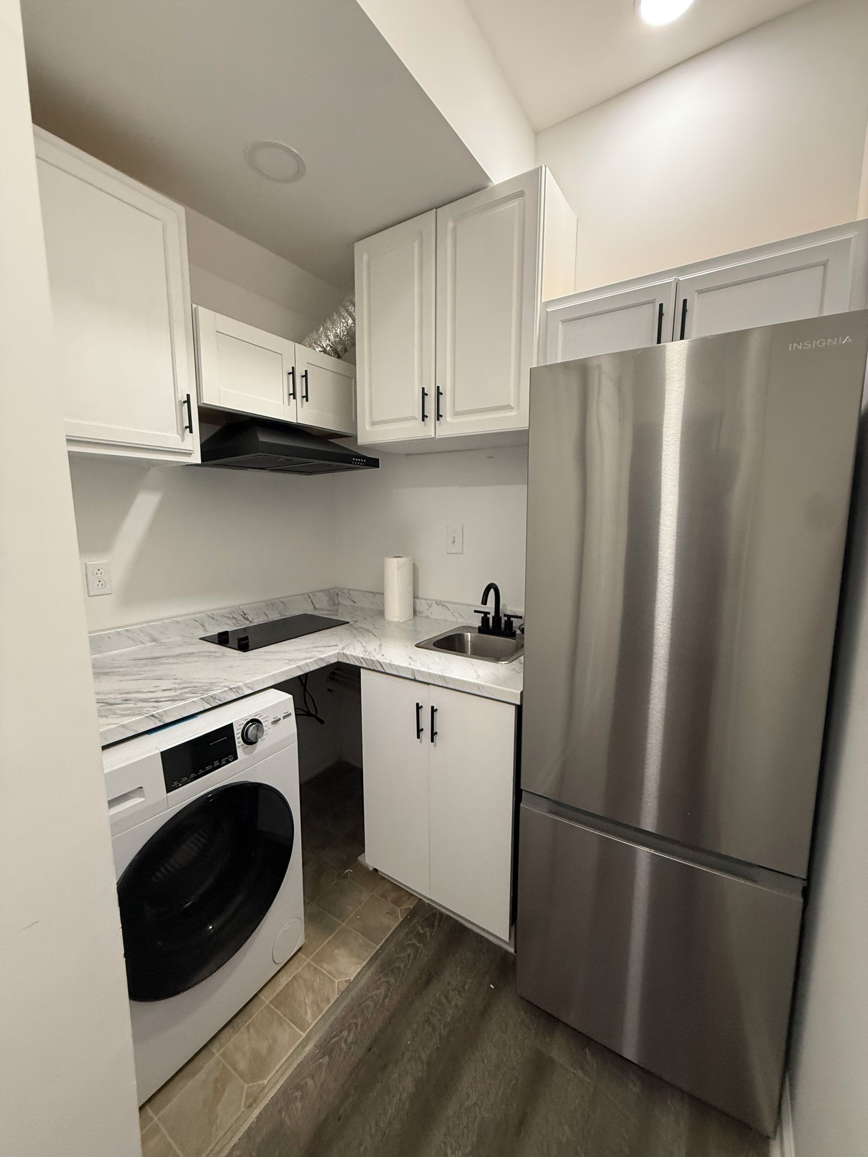 Cocina compacta con muebles blancos, refrigerador de acero inoxidable, lavadora integrada y encimera de mármol.
