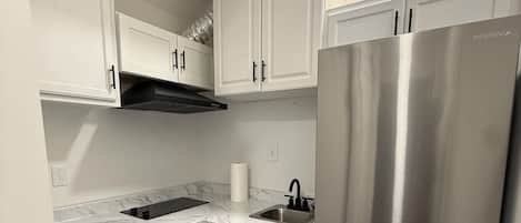 Cocina compacta con muebles blancos, refrigerador de acero inoxidable, lavadora integrada y encimera de mármol.