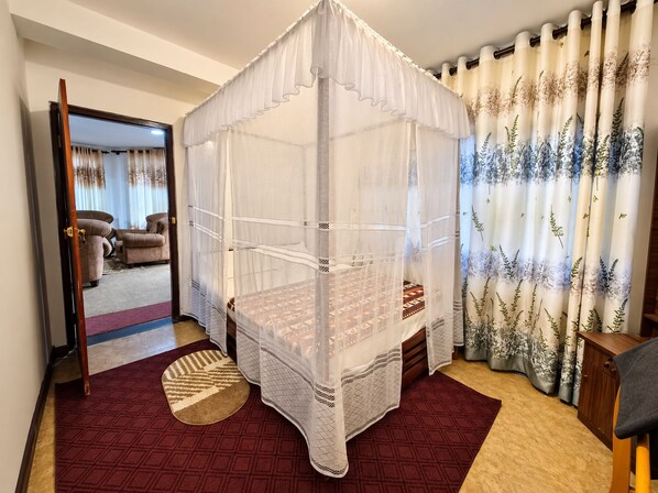 A room with a four-poster canopy bed, a red carpet, and a view of a living area with a sofa and curtains.