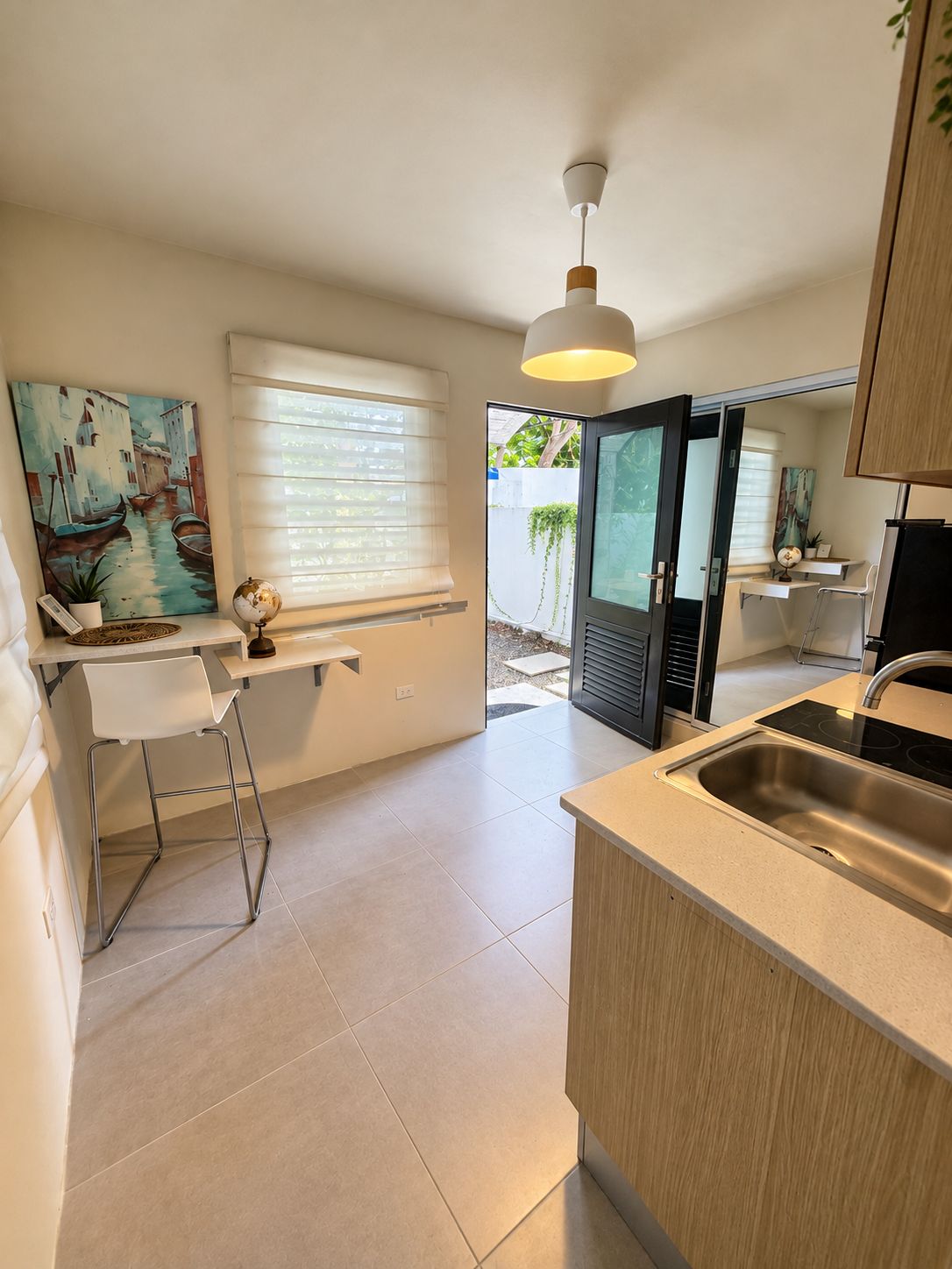 A modern kitchen with a built-in island, a dining area with a white chair, a painting on the wall, and a view through a glass door.