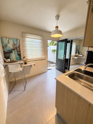 A modern kitchen with a built-in island, a dining area with a white chair, a painting on the wall, and a view through a glass door.