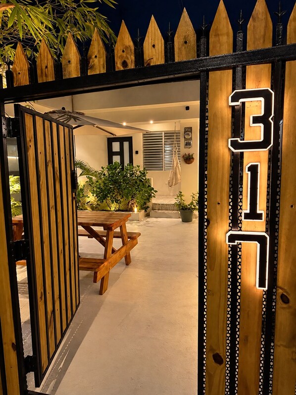 An open wooden gate with a number '317' illuminated on the right side, leading to an outdoor seating area with a table and chairs, surrounded by potted plants.
