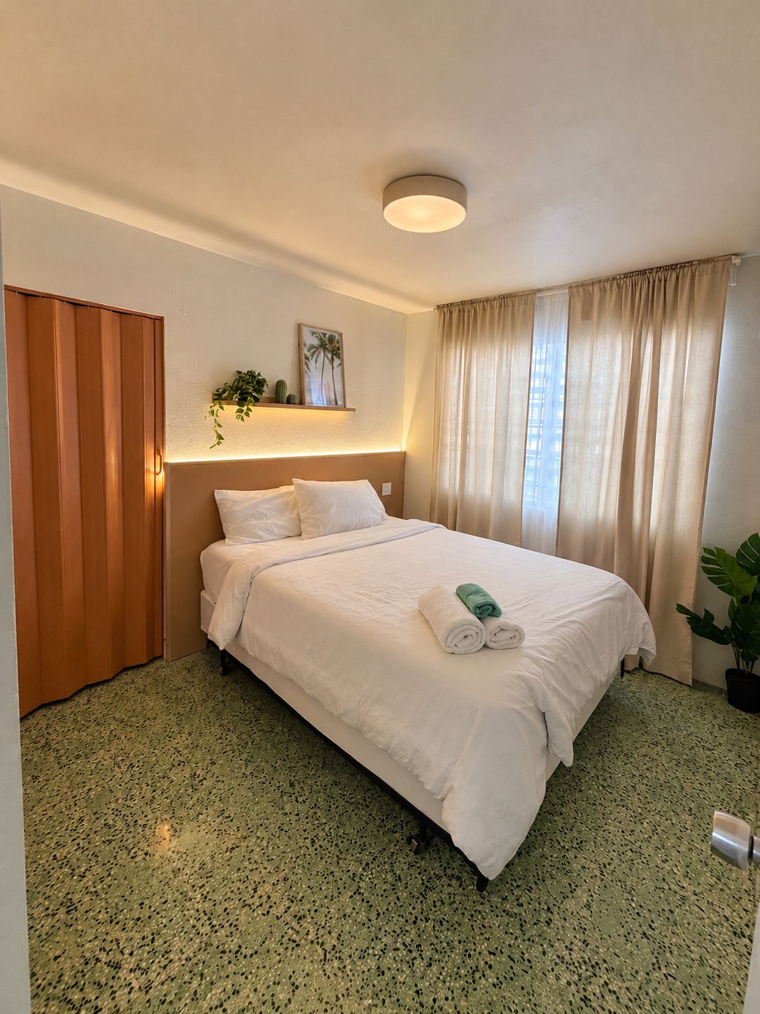 A hotel room with a bed, a bedside table, a wall-mounted shelf with a plant, a window with curtains, and a door.