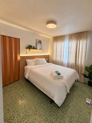 A hotel room with a bed, a bedside table, a wall-mounted shelf with a plant, a window with curtains, and a door.